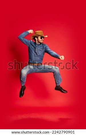 Excited young man in cowboy style outfit having fun, posing isolated over red background. Model in cowboy hat and denim clothes. Fashion, emotions concept. Looks happy, delighted Royalty-Free Stock Photo #2242790901