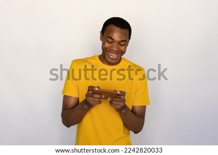 Portrait of cheerful young man surfing internet on mobile phone and laughing. African American guy wearing yellow T-shirt watching video and playing on smartphone. Mobile technology concept