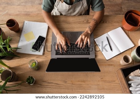 top view of cropped african american florist typing on laptop near copybooks and plants on desk in flower shop