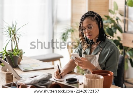 young african american florist in eyeglasses looking at smartphone and writing order in notebook in flower shop Royalty-Free Stock Photo #2242892157