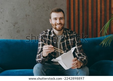 Young man he wears brown shirt eat Chinese food cuisine in takeaway carton container box sitting on blue sofa in own living room apartment stay home indoor flat on weekends. People lifestyle concept