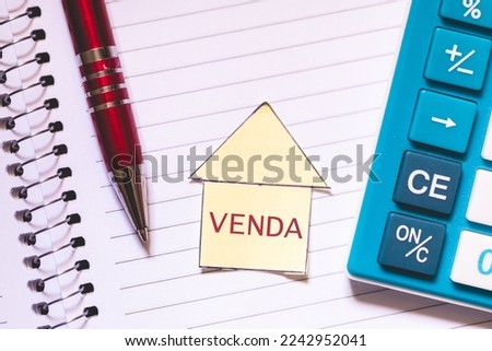 The word sale written in Brazilian Portuguese on a house-shaped piece of paper. A notebook, a calculator and a pen in the composition.
