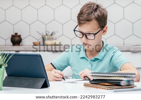 Portrait of preteen serious boy pupil wearing blue T-shirt, glasses, sitting at desk, looking at screen of tablet, doing homework, writing on notebook with left hand. Distant learning, education. Royalty-Free Stock Photo #2242983277