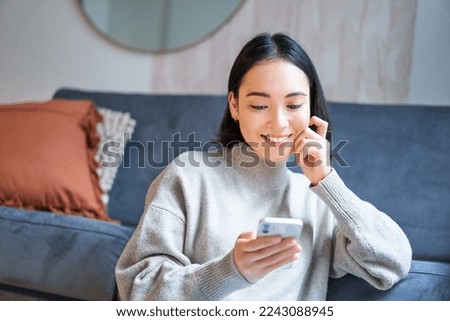 Portrait of smiling asian woman looking intrigued at smartphone screen, interested with smth on mobile phone, sitting coy on floor in living room. Royalty-Free Stock Photo #2243088945