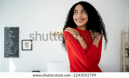 Embrace equity on multiracial Internal Women's Day. Lady African American good mood hands hug herself shoulders enjoy joyful red cloth laundry warmth toothy smile. Royalty-Free Stock Photo #2243092799