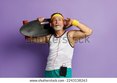 Smiling Man Holding Skateboard. Headphones Senior Man Holding Longboard on Shoulder and Listening Music While Posing Isolated on Violet Background 