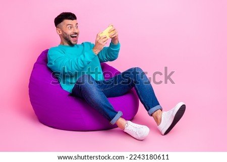 Full size photo of young handsome male sit bean bag play mobile game gamer wear trendy blue garment isolated on pink color background