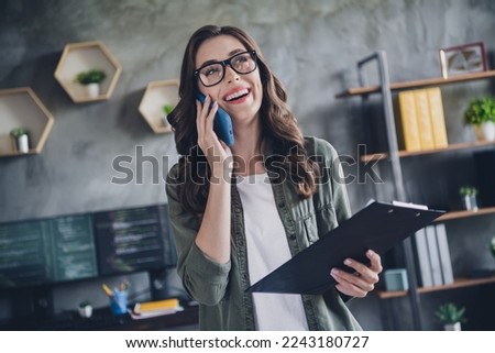 Photo of cheerful positive lady manager wear glasses talking modern gadget laughing holding checklist indoors workstation workshop