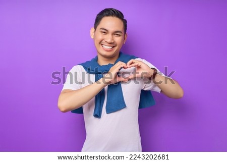 Smiling young Asian man wearing white t-shirt showing shape heart with hands isolated over purple background. people lifestyle concept