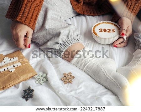 Happy new year 2023 coffee cup with number 2023 over frothy surface in female hand holding while relaxed sitting on the bed with white blanket, gift bag, green wooden snowflake sign. Holidays food art