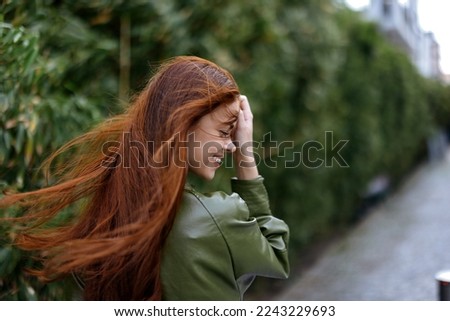 Fashion woman in the city outside posing with a smile against buildings and trees with red long flying hair in the windy weather Royalty-Free Stock Photo #2243229693