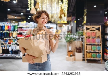 Young woman in casual clothes shopping at supermaket store with craft paper package with groceries using mobile cell phone browsing internet look for recipe inside hypermarket. Gastronomy food concept