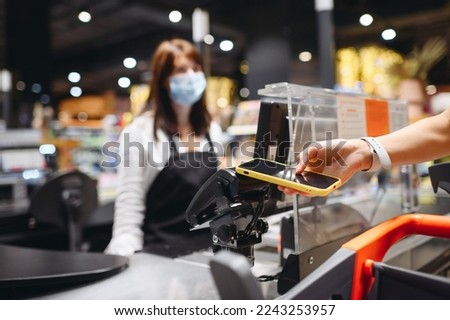 Cropped close up woman hand in casual clothes mask shopping at supermaket with grocery cart put mobile phone to modern bank payment terminal process acquire payments near cashier checkout inside store Royalty-Free Stock Photo #2243253957
