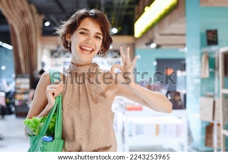 Young smiling happy woman 20s in casual clothes shopping at supermaket store with green eco bag show ok okay gesture produce inside hypermarket. People lifestyle purchasing gastronomy food concept.