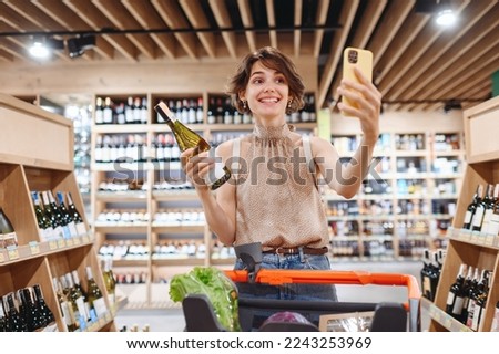 Young happy woman in casual clothes shopping at supermaket store with grocery cart hold white wine alcohol hold bottle do selfie shot on mobile phone inside hypermarket Purchasing gastronomy concept