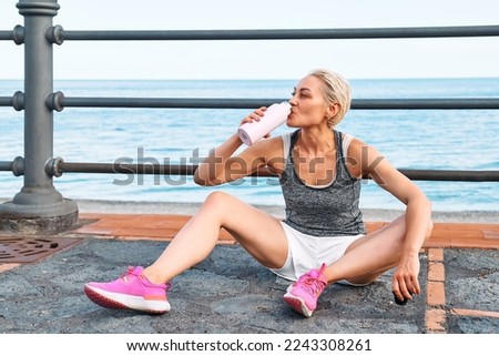 Fit woman relaxes and drinking water on seaside promenade after running and training on a road by the sea. Workout outdoors. Fitness, sport and healthy lifestyle concept. Royalty-Free Stock Photo #2243308261
