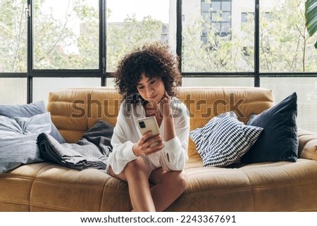 Young african american woman sitting on a sofa in bright loft apartment living room looking cellphone with interest Royalty-Free Stock Photo #2243367691
