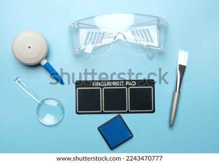 Flatlay picture of toy fingerprint pad, magnifying glass, puffer, brush, safety glass and inkpad on blue background.