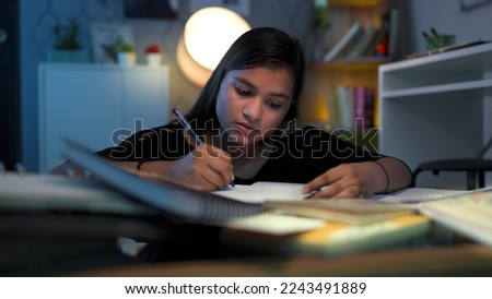Young focused school, college girl with pen in hand write, study, think, doing assignment project homework for exams. Hard working Indian teen concentrating, read alone late hours in night at home  Royalty-Free Stock Photo #2243491889