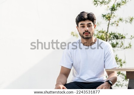 A young handsome bearded man wearing a blank white shirt doing a sitting pose outdoor while facing the cam, mockup shirt image