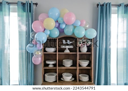 A pastel balloon arch for baby shower decorations with awindows and aqua curtains.
