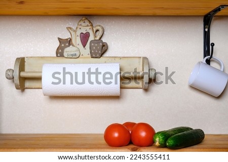 On the kitchen wooden shelf are several red tomatoes and two large green cucumbers. A wooden fixture with a paper towel is installed on the wall. A white mug is hanging on the hook.