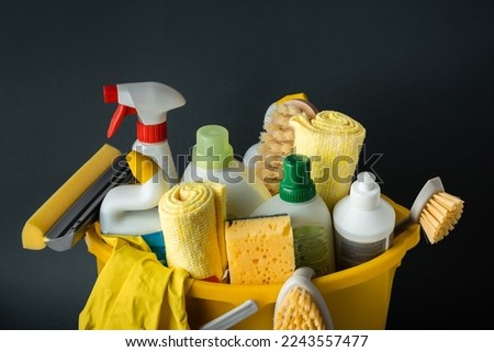 Brushes, bottles with cleaning liquids, sponges, rag and yellow rubber gloves on the black background. Cleaning supplies in the yellow bucket. Cleaning company service advertisement Royalty-Free Stock Photo #2243557477