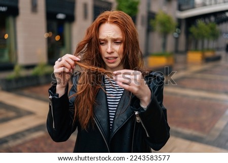 Portrait of displeased pretty young woman touching wet hair after autumn rain standing on beautiful city street. Front view of stressed lady untangling hair after being caught in rain. Royalty-Free Stock Photo #2243583577