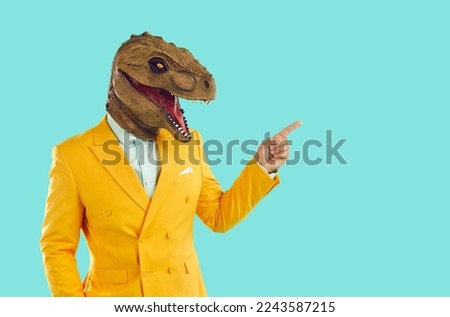 Creative advertising banner. Cool man in trendy rubber dinosaur mask is pointing with his finger at place for advertisement. Man with dinosaur head and in yellow suit isolated on turquoise background.