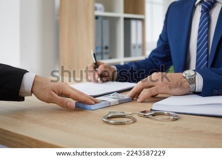 Unrecognizable man in suit gives bribe to authority. Male businessman gives money bundle to man who signs illegal document agreement on office desk with handcuffs. Finance crime, corruption concept Royalty-Free Stock Photo #2243587229