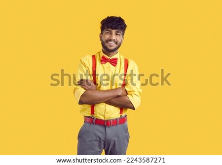 Studio portrait of smiling man in party outfit. Happy young Indian guy in yellow shirt, gray pants, suspenders and red bow tie standing with his arms folded isolated on bright yellow color background