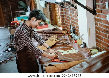 Asian craftsman in apron working with leather making pattern in workshop