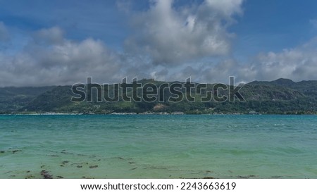 The tropical island is overgrown with green vegetation. Houses are visible near the coast and on the top of the hill. Ripples on the surface of the turquoise ocean. Clouds in the blue sky. Seychelles