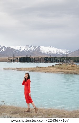 Asian female tourist pose at turquoise color Lake Tekapo with snow capped New Zealand southern alps and Church of Good Shepherd at the background. 