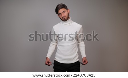 Portrait of frustrated worried brunette man with beard turning out empty pockets showing I have no money gesture, Poor, bankrupt. indoor studio shot isolated on gray background.