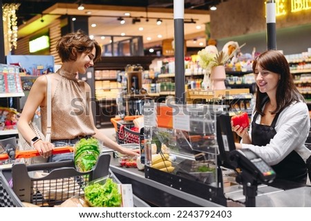 Young cheerful happy woman 20s wear casual clothes shopping at supermaket with grocery cart stand at store checkout pays for groceries cashier inside hypermarket Purchasing gastronomy food concept.