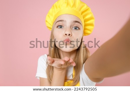 Close up fun teen girl chef cook confectioner housewife baker in yellow apron white t-shirt cap do selfie shot on mobile phone blowing air kiss isolated on pink background studio. Food cake concept