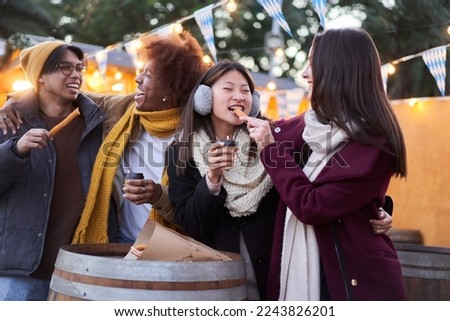 Happy friends eating chocolate with churros street food at the city in the night outdoors. Tourist group of four people having fun in winter time. One girl feeds the other. Friendship concept.