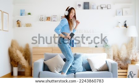 On the sofa in her living room, a young Asian woman dances. Asian woman with a smile relaxing at home, happy and in good mental and physical health, wearing headset and listening to music Royalty-Free Stock Photo #2243860783