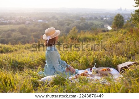 Rear view of woman in a long summer dress and straw hat with short hair sitting on a white blanket with fruits and pastries. Concept of having picnic in a city park during summer holidays or weekends.