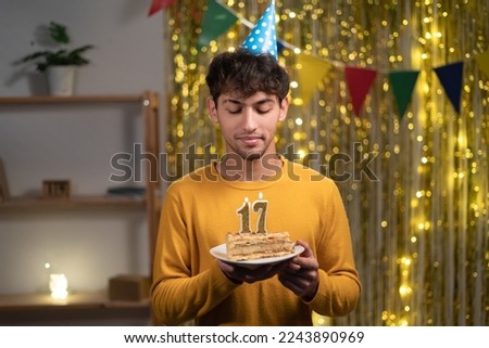 Young man in cone party hat to make wish holding his birthday cake with candles number 17 during celebration at home. Seventeens birthday concept Royalty-Free Stock Photo #2243890969