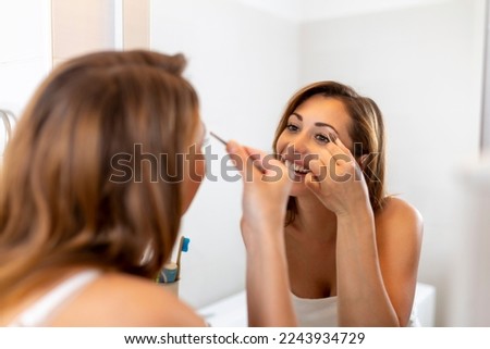Self care and make up concept. Waist up portrait of young careful woman in white shirt plucking her eyebrows by tweezers before mirror in bathroom