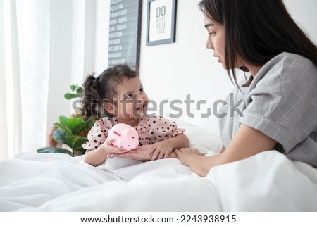 Happy cute Asian little daughter smiling and money saving teach from mom, sitting on bed, speaking, telling funny story. Saving money with pink piggy bank. Education and learning concept