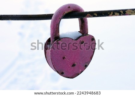 an old pink lock in the shape of a heart covered with snow hangs on an iron railing