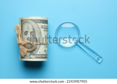 Flatlay picture of role fake cash and magnifying glass on blue background. Finding financial solution concept.
