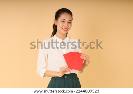 Portrait of a pretty cheerful Vietnamese young lady in office outfit, with hand gesture and cute face expression, hold lucky money red envelop bag. Concept of Tet Holiday (Lunar New Year)