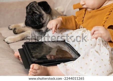 A one-year-old girl with a pug watches cartoons on a tablet sitting on the couch. Gadgets for kids, screen time.