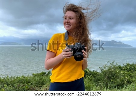 Portrait of happy positive girl traveller, young beautiful woman photographer with camera in hand taking a picture, photo of sea, ocean landscape. Traveling with camera in tropical country