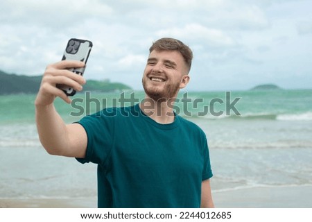 Young guy, handsome happy man at sea is taking picture of hisself, selfie at camera of his phone, using smartphone for social media at summer beach in tropical exotic country. Blogger, vlog