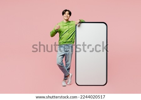 Full body happy young man of Asian ethnicity wear green hoody big huge blank screen mobile cell phone smartphone with area show thumb up gesture isolated on plain pastel light pink background studio